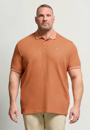 Stämmiger Mann mit kurzen Haaren und Bart, der ein orangefarbenes kurzärmeliges Polohemd und hellbeige Hose trägt und vor einem einfarbigen Hintergrund steht.