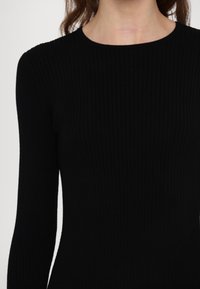 Femme portant un haut noir côtelé ajusté à manches longues avec un col rond, raccourci au niveau du cou et du haut du torse.