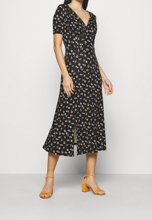 Kvinde iført en sort blomsterprintet midi-kjole med korte ærmer og frontslids, kombineret med sennepsgule blokhælte sandaler.