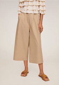 Pantalons larges beiges en coton, avec des plis à l'avant et une taille haute. Associés à des sandales en cuir tressé marron.
