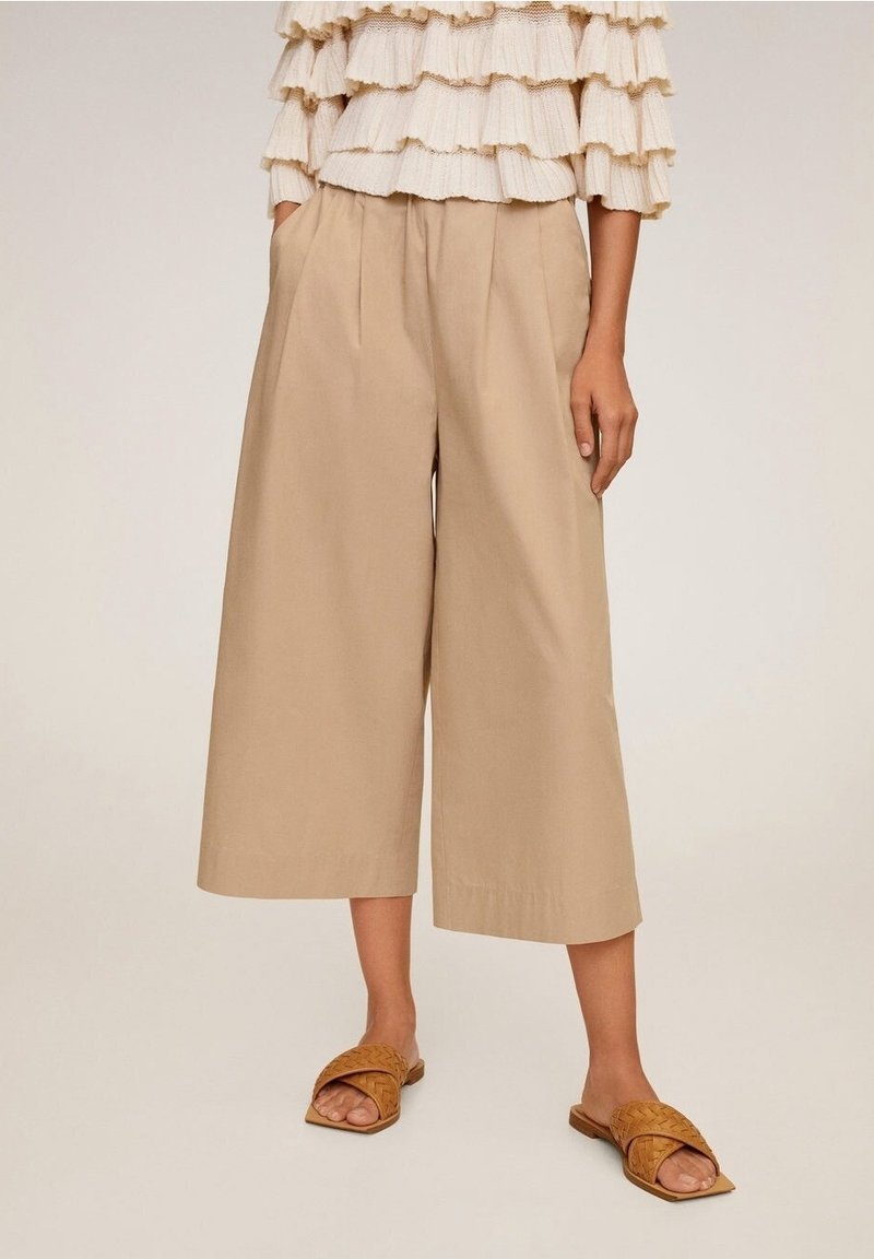 Pantalons larges beiges en coton, avec des plis à l'avant et une taille haute. Associés à des sandales en cuir tressé marron.