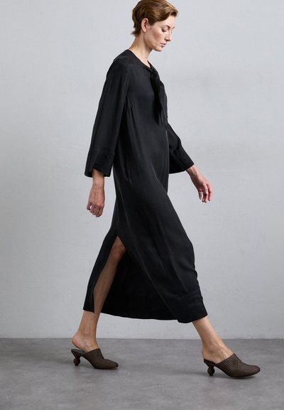 Black long-sleeve dress with side slits, made of a smooth fabric. Paired with brown woven mules featuring rounded heels.