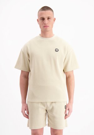 Beige wafelgebreide T-shirt met korte mouwen en een ronde hals, voorzien van een klein zwart logo op de borst, gecombineerd met bijpassende shorts. Minimalistisch ontwerp.