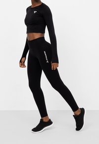 Černý dlouhý rukávový crop top a legíny s vysokým pasem, vyrobené z elastického materiálu s detailem textu "SQUATPROOF", doplněné o černé sportovní boty.