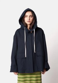 Marineblaue Kapuzenjacke mit lockerem Schnitt, ausgestattet mit zwei frontalen Taschen, einer Knopfverschluss und beigen Kordeln. Strukturierter Stoff mit weiten Ärmeln.