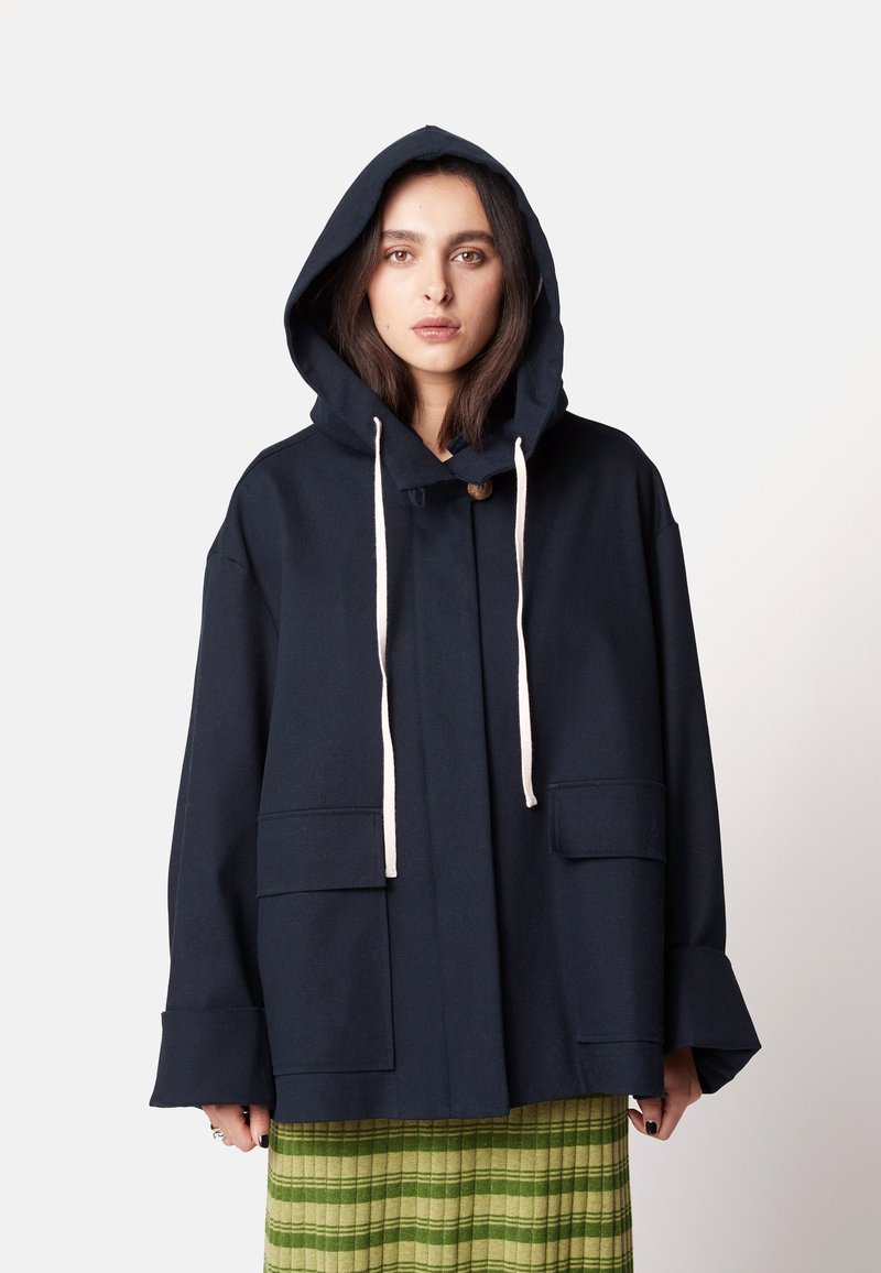 Marineblaue Kapuzenjacke mit lockerem Schnitt, ausgestattet mit zwei frontalen Taschen, einer Knopfverschluss und beigen Kordeln. Strukturierter Stoff mit weiten Ärmeln.