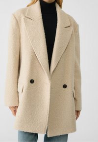 Cream-colored double-breasted coat made of textured fabric, featuring large black buttons and notched lapels. Worn over a black turtleneck.