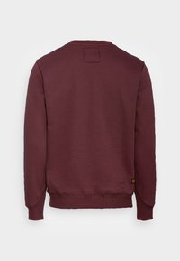 Sudadera burdeos con cuello redondo, puños y dobladillo acanalados. Textura suave, con un sutil detalle de parche en la parte posterior.