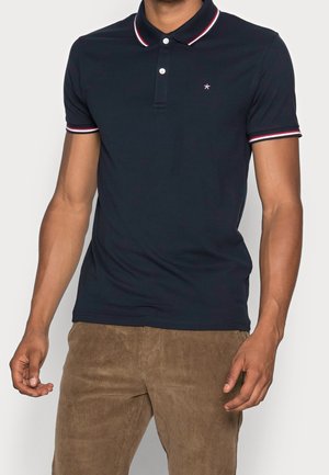 Mann trägt ein marineblaues Poloshirt mit rot-weiß gestreiftem Kragen und Ärmeln, kombiniert mit braunen Cordhosen.