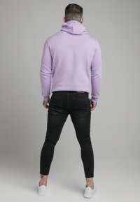 Sudadera morada con cordón, mangas largas y puños acanalados. Jeans negros ajustados, textura de mezclilla y zapatillas blancas de contraste.