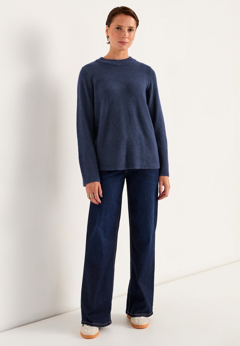 Marineblauer Strickpullover mit Rundhalsausschnitt und geripptem Saum, kombiniert mit weit geschnittenen dunkelblauen Jeans und weißen Sneakers mit sandfarbenen Sohlen.