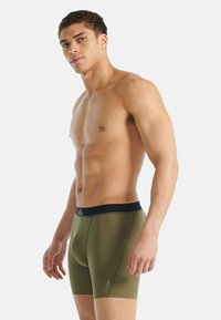 Muž bez trička v olivově zelených boxerkách s černým pasem, stojící bokem na bílém pozadí.