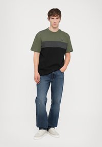 Tricou grafic cu blocuri de culoare olive, gri și negru; cu guler rotund; purtat cu blugi decolorati și teniși albi.
