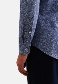 Camicia a maniche corte blu con motivo floreale, realizzata in cotone texturizzato, con piccoli motivi bianchi e gialli e bottoni bianchi sui polsini.