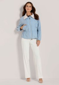 Lichtblauwe cropped jas met een witte kraag, ritssluiting en elastische tailleband; heeft twee zijzakken en lange mouwen.