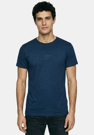 Jeune homme aux cheveux bouclés et foncés portant un t-shirt bleu marine Lee Cooper et un jean noir, debout devant un fond clair uni.