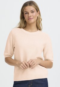 Gaiši rozā īss piedurkņu adīts džemperis ar ribotu apaļu kakla izgriezumu un gludu tekstūru, kombinēts ar tumši ziliem džinsa džinsiem.