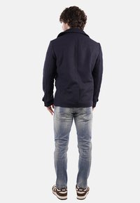 Giacca in misto lana blu navy con ampio colletto, spalle strutturate e una texture liscia, abbinata a jeans in denim chiaro e sneakers marroni.