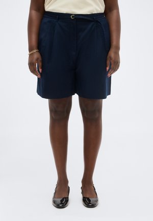 Osoba w granatowych szortach z paskiem, beżowym topie, czarnych błyszczących balerinach i złotej bransoletce, stojąca na tle jednolitego białego tła.