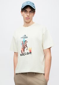 Εκρού κοντομάνικη μπλούζα με πολύχρωμο γραφικό ενός καουμπόη, αλόγου και UFO· περιλαμβάνει τη λέξη "VOLCOM" με έντονα μαύρα γράμματα.