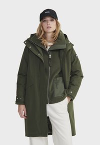Manteau imperméable vert avec capuchon, fermeture éclair à l'avant et poches latérales. Couche intérieure en polaire visible, associé à une casquette noire. Pantalons blancs.
