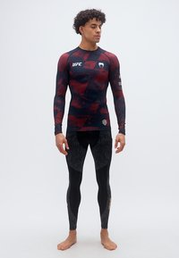 Άνδρας όρθιος, ξυπόλητος, φορώντας μακρυμάνικο μαύρο και κόκκινο συμπιεστικό μπλουζάκι UFC και μαύρα συμπιεστικά κολάν με σχέδια.