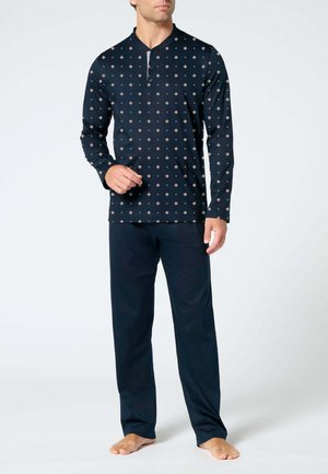 Uomo che indossa un pigiama blu navy a due pezzi con maglia a maniche lunghe fantasia e pantaloni tinta unita, in piedi a piedi nudi su sfondo neutro.