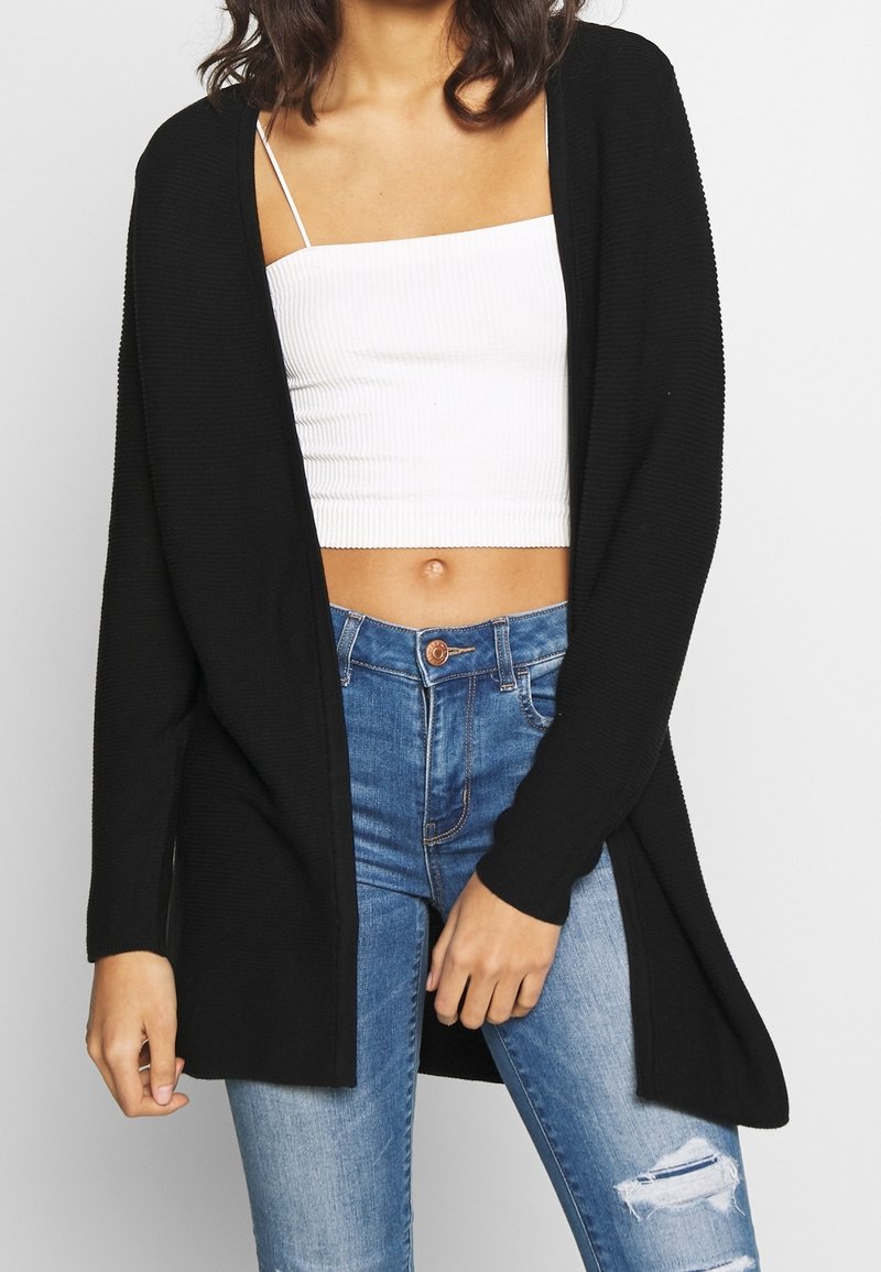 Svart cardigan med öppen front, långa ärmar och ribbat mönster, bärs över en vit kortärmad topp och blå slitna jeans.