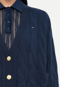 Cárdigan de punto trenzado en azul marino con botones dorados, pequeño logo en rojo-blanco-azul, llevado sobre una camisa de encaje azul marino con botones y cuello.
