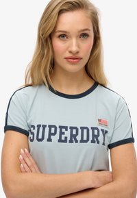 T-shirt bleu clair avec des accents marine, arborant le texte en gras "SUPERDRY" et une petite patch de drapeau américain sur la poitrine. Tissu doux, design ajusté.