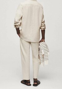 Beżowa lniana koszula i spodnie o luźnym kroju, z podwiniętymi rękawami. Trzyma biały sweter w paski. Na stopach czarne sandały.