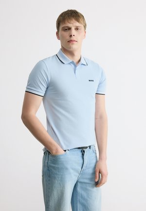 Mladý muž v lehce modré polo košili s krátkými rukávy a logem "BOSS" a v lehce modrých džínách stojí před jednobarevným pozadím.