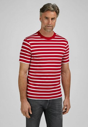 Mann mit grauem Haar und Bart, trägt ein rot-weiß gestreiftes Kurzarmhemd und dunkelgraue Jeans, steht vor einfachem Hintergrund.