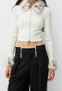 White cropped zip-up top with ribbed texture and faux fur collar, featuring black pants with frayed edges and fur accents on cuffs.