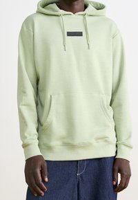 Felpa con cappuccio verde chiaro con tasca a marsupio, cappuccio con coulisse regolabile e patch logo nero sul petto. Tessuto morbido e polsini a costine.