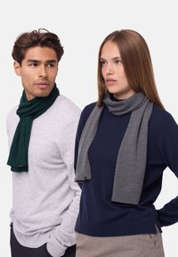 Grønne og grå strikkede tørklæder hænger om halsen på to modeller. Det grønne tørklæde har en ribbet struktur, mens det grå tørklæde har en glat finish.