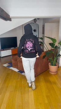 Fekete kapucnis pulóver rózsaszín virágmintával és felirattal a hátán, világosszürke melegítőnadrággal és fekete cipővel, fa padlón.