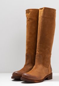 Un paio di alti stivali in suede marrone con punte arrotondate e tacchi in legno a blocco su una superficie bianca contro uno sfondo semplice.