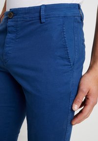 Modré bavlněné kalhoty s hladkou texturou, vybavené zapínáním na knoflík, poutky na opasek a bočními kapsami. Detailní pohled na látku a střih.