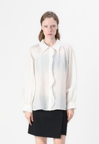 Blusa bianca a maniche lunghe in tessuto strutturato, con chiusura frontale a bottoni, dettaglio arricciato e colletto classico. Abbinata a una gonna nera.