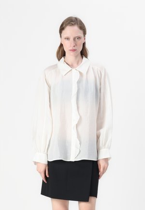 Blusa bianca a maniche lunghe in tessuto strutturato, con chiusura frontale a bottoni, dettaglio arricciato e colletto classico. Abbinata a una gonna nera.