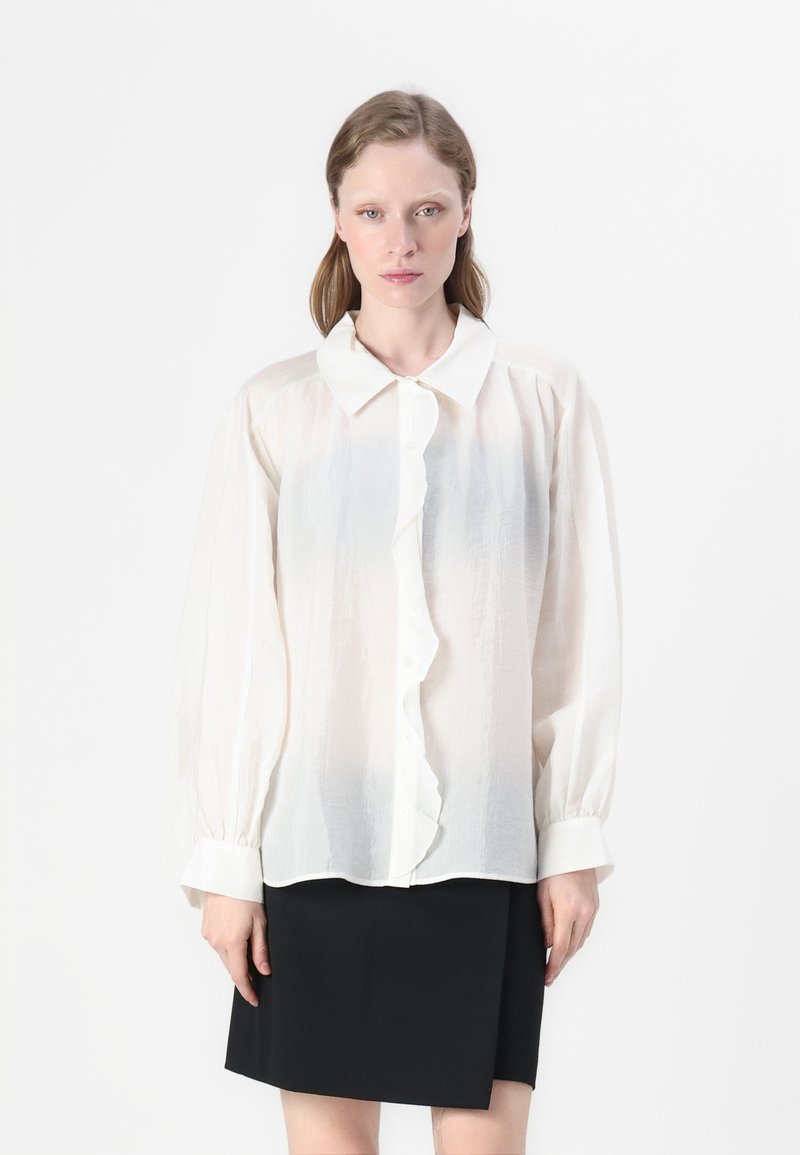 Blusa bianca a maniche lunghe in tessuto strutturato, con chiusura frontale a bottoni, dettaglio arricciato e colletto classico. Abbinata a una gonna nera.