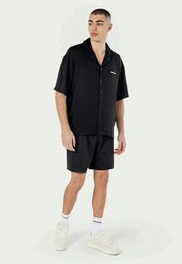 Camisa preta de manga curta com botões e gola, combinada com calções de malha pretos e ténis brancos. Design simples, sem padrões ou detalhes.
