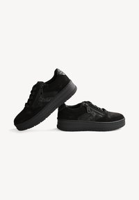 Paire de baskets noires basses avec semelles épaisses, fermetures éclair latérales et lacets à l'avant, présentée sur un fond clair uni.