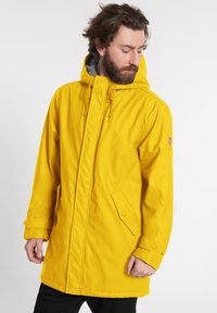 Parka gialla impermeabile con cappuccio regolabile, due tasche frontali e una morbida fodera grigia; presenta una vestibilità comoda e ampia.