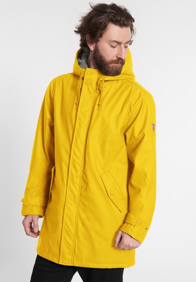 Parka gialla impermeabile con cappuccio regolabile, due tasche frontali e una morbida fodera grigia; presenta una vestibilità comoda e ampia.