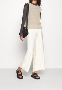 Femme portant un haut noir transparent à manches longues, un gilet beige en tricot, un pantalon large blanc, des sandales noires à boucles, et portant un sac à bandoulière noir.