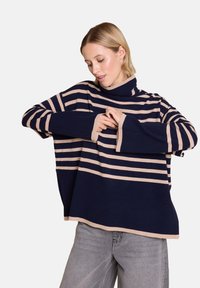 Marineblå og beige stripete strikkegenser med høy hals, løs passform og brede ermer. Har kontrasterende mansjetter og nederkant.