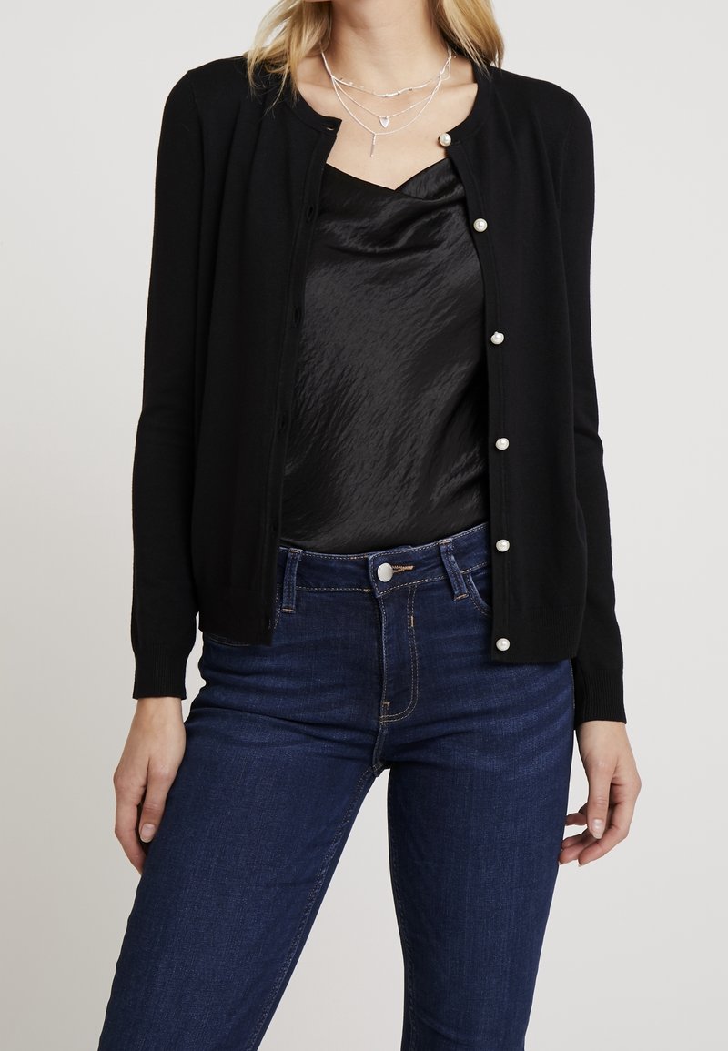 Femme portant un cardigan noir avec des boutons en perles sur un caraco en satin noir et un jean bleu foncé, accessoirisée avec des colliers argentés superposés.