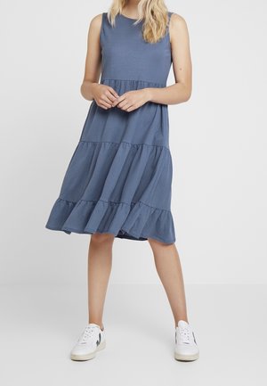 Kobieta w niebieskiej, bezrękawnikowej, wielowarstwowej sukience do kolan i białych trampkach, stojąca na tle jasnego, jednolitego tła.